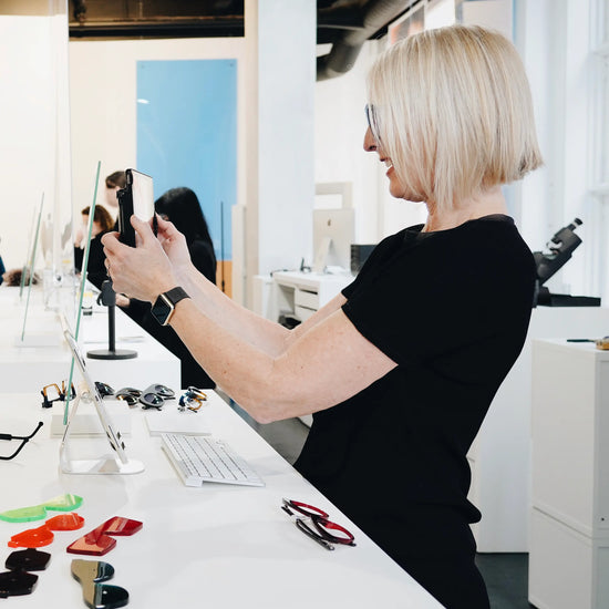 Opticienne dans une boutique de lunettes à Montréal prenant une photo d’une cliente pendant un examen de la vue, devant un comptoir rempli de montures colorées.